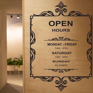 May include: Black and white sign with ornate frame on a wood background. The sign reads "Open Hours" with the following hours: Monday - Friday 7AM - 5PM, Saturday 7AM - 9PM, Sunday Closed.