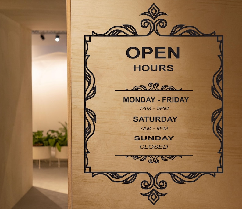 May include: Black and white sign with ornate frame on a wood background. The sign reads "Open Hours" with the following hours: Monday - Friday 7AM - 5PM, Saturday 7AM - 9PM, Sunday Closed.