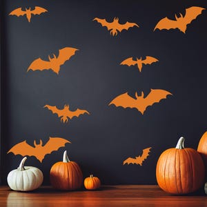 Może przedstawiać: Pomarańczowe naklejki ścienne z nietoperzami zdobią ciemną ścianę, tworząc motyw Halloween. Kilka dyni o r&oacute;żnych rozmiarach, w tym jedna biała, jest ułożonych na podłodze. Scena przywołuje świąteczną jesienną atmosferę.