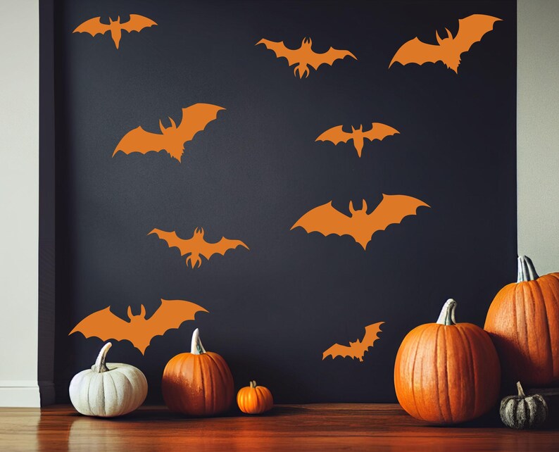 Może przedstawiać: Pomarańczowe naklejki ścienne z nietoperzami zdobią ciemną ścianę, tworząc motyw Halloween. Kilka dyni o r&oacute;żnych rozmiarach, w tym jedna biała, jest ułożonych na podłodze. Scena przywołuje świąteczną jesienną atmosferę.