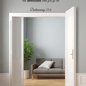 Pu&ograve; includere: Una porta bianca con una maniglia nera &egrave; aperta per rivelare un soggiorno con un divano grigio, una pianta verde e un pavimento in legno. La parete dietro la porta ha una citazione di testo nero che recita "You will be Blessed when you come in and Blessed when you go out Deuteronomy 28:6".