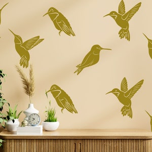 May include: A beige wall with nine gold hummingbird silhouettes. The wall is decorated with a wooden shelf with a clock, a vase with dried grass, and two potted plants.