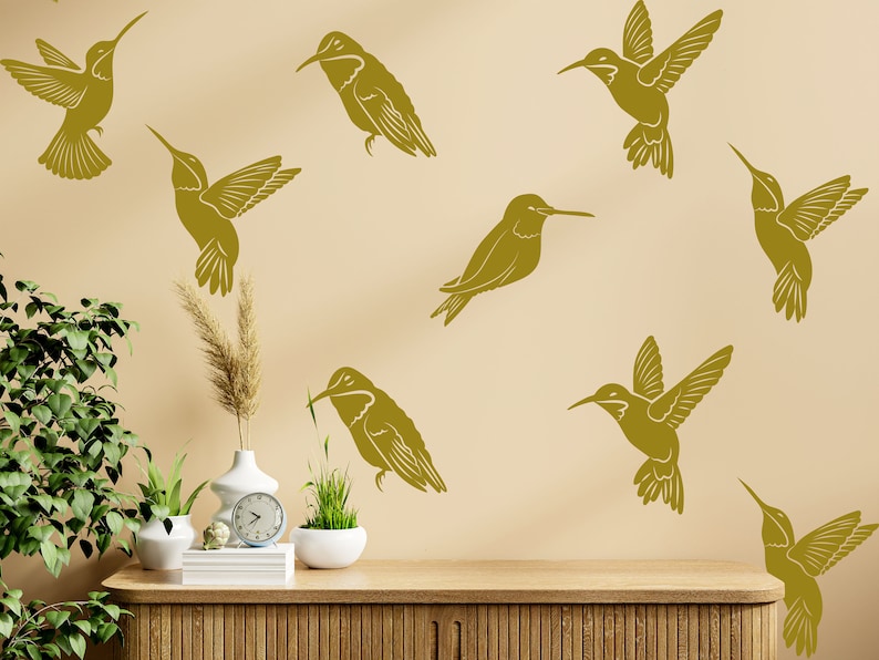 May include: A beige wall with nine gold hummingbird silhouettes. The wall is decorated with a wooden shelf with a clock, a vase with dried grass, and two potted plants.