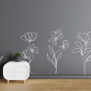 May include: White line art floral wall decals. Three different floral designs are displayed on a dark grey wall. The decals are simple, elegant, and modern. The room also features a white bedside table, a grey chair, and a small potted plant.