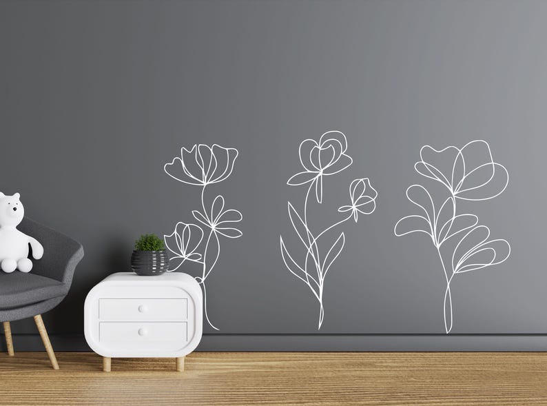 May include: White line art floral wall decals. Three different floral designs are displayed on a dark grey wall. The decals are simple, elegant, and modern. The room also features a white bedside table, a grey chair, and a small potted plant.