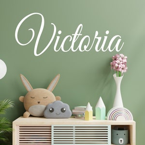 May include: A green wall with the name "Victoria" written in white cursive lettering. A wooden shelf with a rainbow toy, a vase of pink flowers, a cloud-shaped toy, a bunny-shaped toy, and a stack of books.