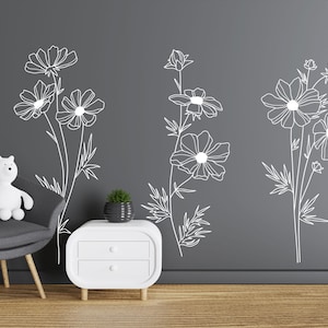 K&ouml;nnte beinhalten: Drei verschiedene Wandaufkleber mit floralen Motiven in wei&szlig;er Umrandung. Die Aufkleber befinden sich an einer grauen Wand mit einem wei&szlig;en Beistelltisch und einem grauen Sessel.