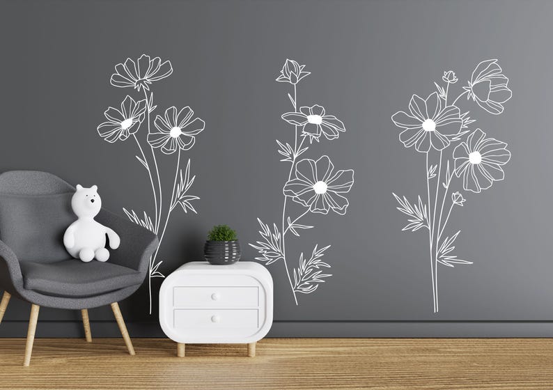 K&ouml;nnte beinhalten: Drei verschiedene Wandaufkleber mit floralen Motiven in wei&szlig;er Umrandung. Die Aufkleber befinden sich an einer grauen Wand mit einem wei&szlig;en Beistelltisch und einem grauen Sessel.