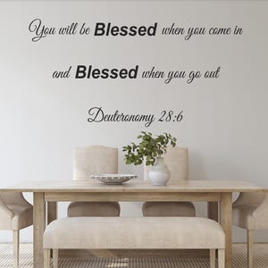 Pu&ograve; includere: Un adesivo da parete con la citazione "You will be Blessed when you come in and Blessed when you go out Deuteronomy 28:6" in corsivo nero. L'adesivo &egrave; esposto su una parete bianca sopra un tavolo da pranzo in legno con due sedie e una panca.