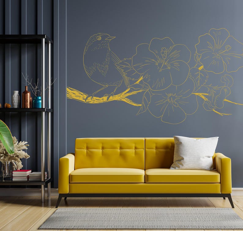 May include: A yellow sofa with a white pillow sits in front of a gray wall with a gold bird and flower design.