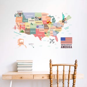 May include: A colorful map of the United States with state names and landmarks. The map is on a white wall above a wooden desk with a stack of books and a wooden chair. The American flag is displayed next to the map.