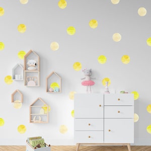 May include: A white dresser with six drawers and wooden knobs sits in a room with a light gray wall decorated with yellow watercolor polka dots. A toy cat in a pink tutu sits on top of the dresser. A toy wagon with a cat face and wheels is in the foreground.