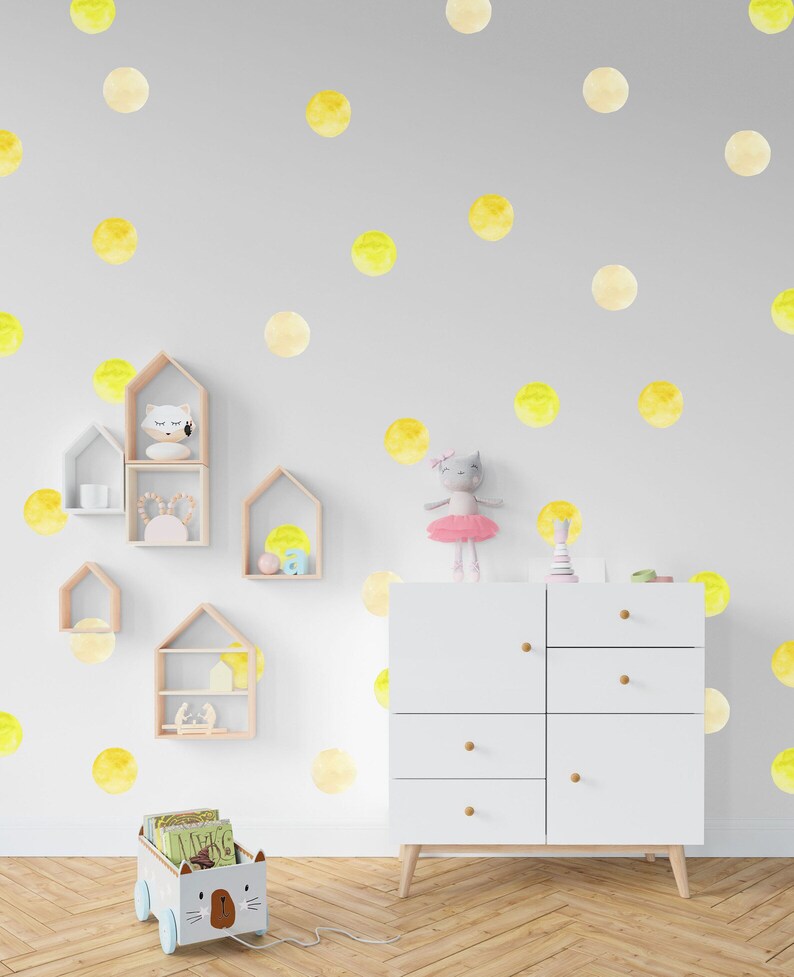 May include: A white dresser with six drawers and wooden knobs sits in a room with a light gray wall decorated with yellow watercolor polka dots. A toy cat in a pink tutu sits on top of the dresser. A toy wagon with a cat face and wheels is in the foreground.