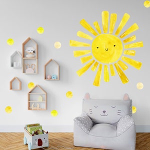 May include: A yellow sun with a smiling face and yellow polka dots on a white wall. A gray cat-shaped chair with a white polka dot pattern is in the foreground. There is a wooden toy cart with books and a cat face on the front.