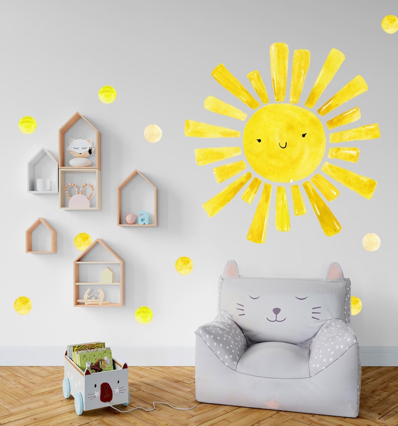 May include: A yellow sun with a smiling face and yellow polka dots on a white wall. A gray cat-shaped chair with a white polka dot pattern is in the foreground. There is a wooden toy cart with books and a cat face on the front.