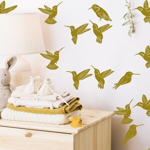 May include: A white wall with a nursery theme featuring gold hummingbird decals. A wooden dresser with a stuffed bunny, folded clothes, and a pacifier on top.