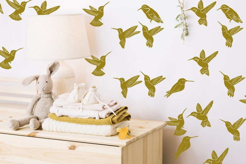 May include: A white wall with a nursery theme featuring gold hummingbird decals. A wooden dresser with a stuffed bunny, folded clothes, and a pacifier on top.
