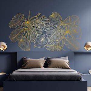 Op de afbeelding: Een moderne slaapkamer met een marineblauwe muur met een gouden bladeren design. Het bed heeft bruine kussens en een grijze sprei. Twee hanglampen hangen aan het plafond. Een kleine plant en een klok staan op een nachtkastje.