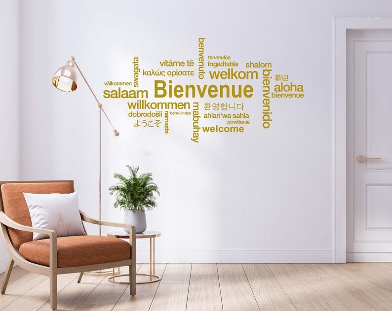 May include: A white wall with a gold word cloud of welcome greetings in different languages, including English, French, Spanish, German, Italian, Japanese, and Arabic. The word cloud is centered on the wall and is surrounded by a white door and a brown armchair with a white pillow.