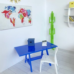 以下が含まれることがあります： 青い机に白い椅子と青い箱が置かれている。机の隣には緑色のサボテンの植物が立っている。机の後ろの壁には世界地図が掛かっている。