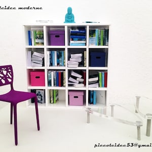May include: A white bookshelf with 12 compartments, filled with books and colorful storage boxes. A purple chair with a unique design sits in front of the bookshelf. A clear glass table with silver legs sits in the foreground.