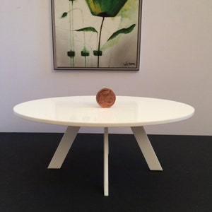 Puede incluir: Mesa redonda blanca con tres patas. La mesa está hecha de madera y tiene un acabado blanco. Una pequeña moneda está encima de la mesa.