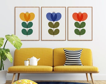 Puede incluir: Tres impresiones enmarcadas de flores estilizadas con pétalos de diferentes colores. Las flores son amarillas, azules y rojas. Las impresiones están colgadas en una pared blanca sobre un sofá amarillo con un cojín a rayas.