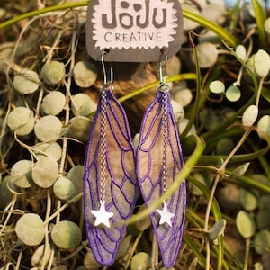 Puede incluir: Pendientes de alas de hada con ribete morado y una cadena plateada con un dije de estrella blanca. Los pendientes cuelgan de una etiqueta con el texto "Joju Creative". El fondo es una mezcla de plantas verdes.