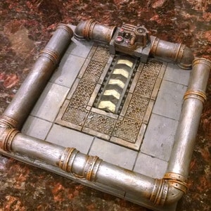 May include: A silver and brown metal model of a sewer grate with a control panel and a striped track. The model is set on a brown surface.