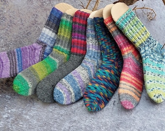 May include: A collection of seven colorful knitted socks, each with a unique pattern and color scheme. The socks are arranged in a row, showcasing a variety of hues including purple, green, blue, red, and orange. The socks are displayed on a neutral gray surface.