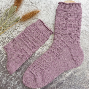 May include: A pair of lavender-colored knitted socks with a textured pattern. One sock is fully extended, while the other is folded down. The socks are displayed on a light gray surface, with dried plants in the background.
