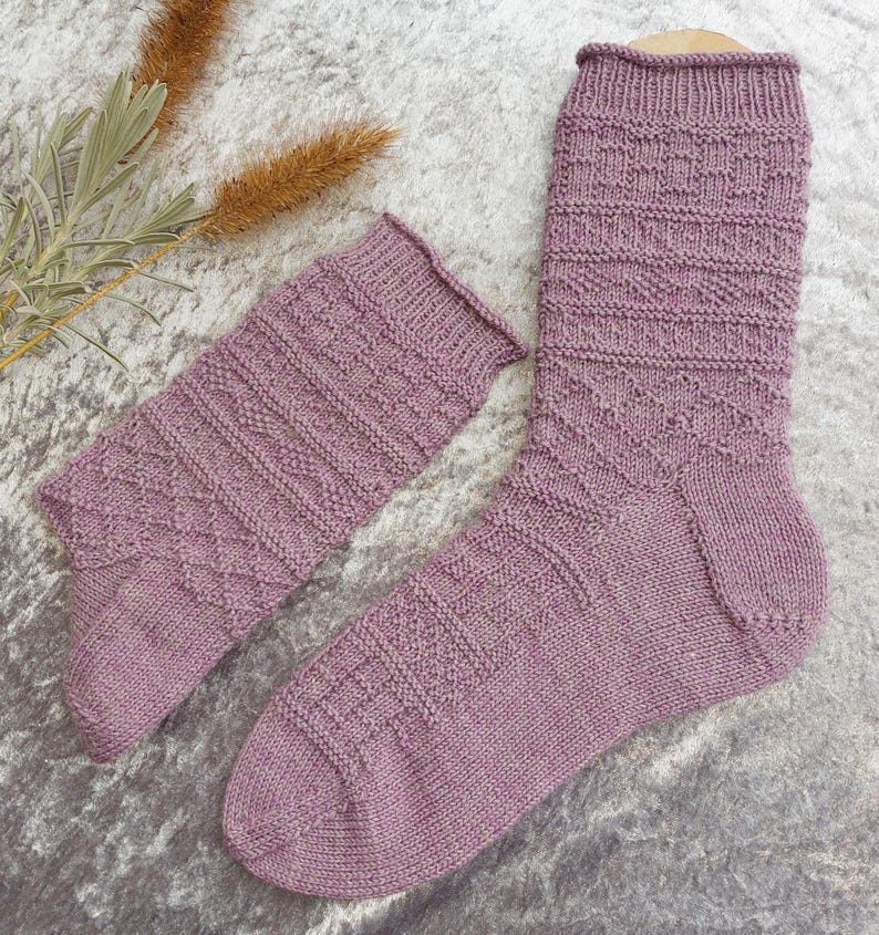 May include: A pair of lavender-colored knitted socks with a textured pattern. One sock is fully extended, while the other is folded down. The socks are displayed on a light gray surface, with dried plants in the background.