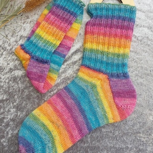 May include: A pair of colorful, hand-knitted socks with a rainbow striped pattern. The socks feature a vibrant mix of colors, including blue, green, yellow, orange, pink, and purple. The socks are displayed on a gray surface.