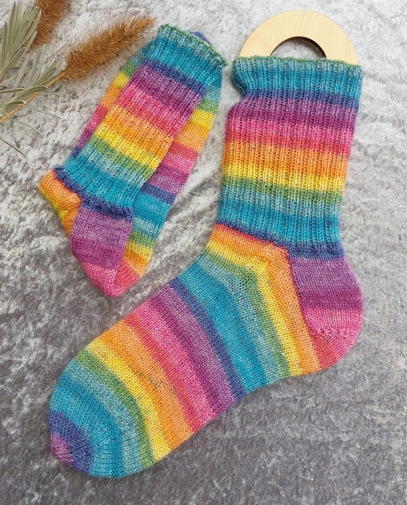 May include: A pair of colorful, hand-knitted socks with a rainbow striped pattern. The socks feature a vibrant mix of colors, including blue, green, yellow, orange, pink, and purple. The socks are displayed on a gray surface.