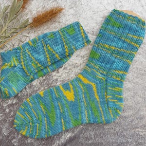 May include: A pair of hand-knitted socks in shades of blue, green, and yellow. The socks feature a striped pattern with a mix of colors. The socks are made of a soft, textured yarn. One sock is larger than the other, suggesting a matching pair.