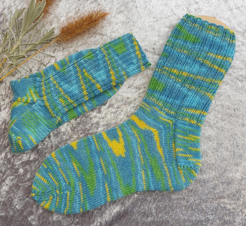 May include: A pair of hand-knitted socks in shades of blue, green, and yellow. The socks feature a striped pattern with a mix of colors. The socks are made of a soft, textured yarn. One sock is larger than the other, suggesting a matching pair.