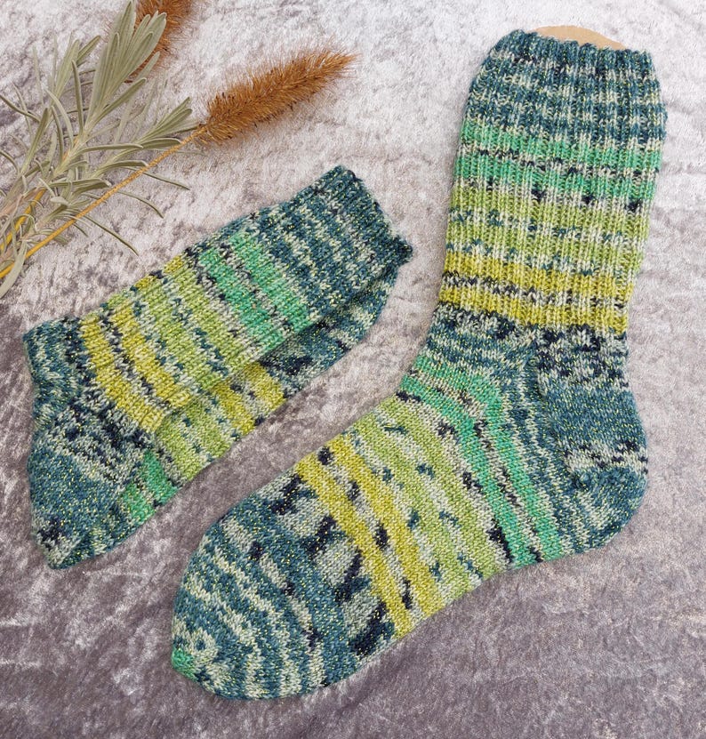 May include: A pair of hand-knitted socks in shades of green, blue, and yellow. The socks have a textured pattern and are displayed on a gray surface. The color gradient creates a visually appealing design, perfect for cozy wear.