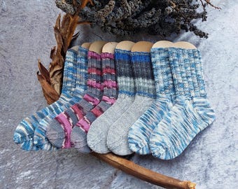 May include: A collection of four pairs of hand-knitted socks displayed in a row. The socks feature various color patterns including blue and white stripes, magenta and grey variegated tones, solid grey with blue bands, and a blue and white speckled design.