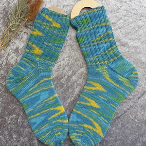 May include: A pair of hand-knitted socks with a blue, yellow, and green striped pattern. The socks are displayed on wooden sock forms. The color scheme is a mix of blues, yellows, and greens, creating a vibrant and textured appearance.