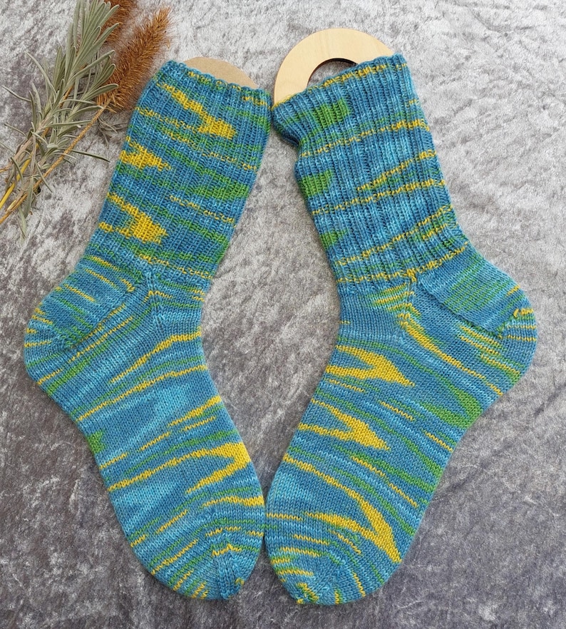 May include: A pair of hand-knitted socks with a blue, yellow, and green striped pattern. The socks are displayed on wooden sock forms. The color scheme is a mix of blues, yellows, and greens, creating a vibrant and textured appearance.