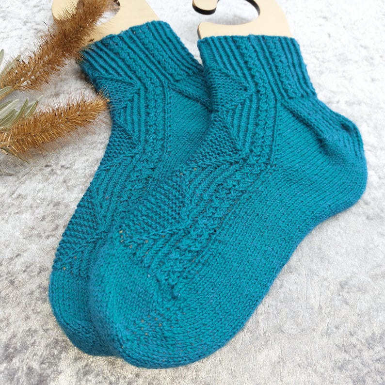 May include: A pair of teal-colored knitted socks with a cable knit design. The socks are displayed on wooden hangers against a light background. The socks have a ribbed cuff and a textured pattern running down the front.