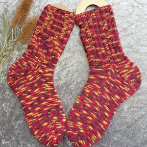May include: A pair of colorful knitted socks. The socks are primarily red with yellow, blue, and green accents. The socks are displayed on wooden sock forms. The socks have a ribbed cuff and a patterned design throughout the body.