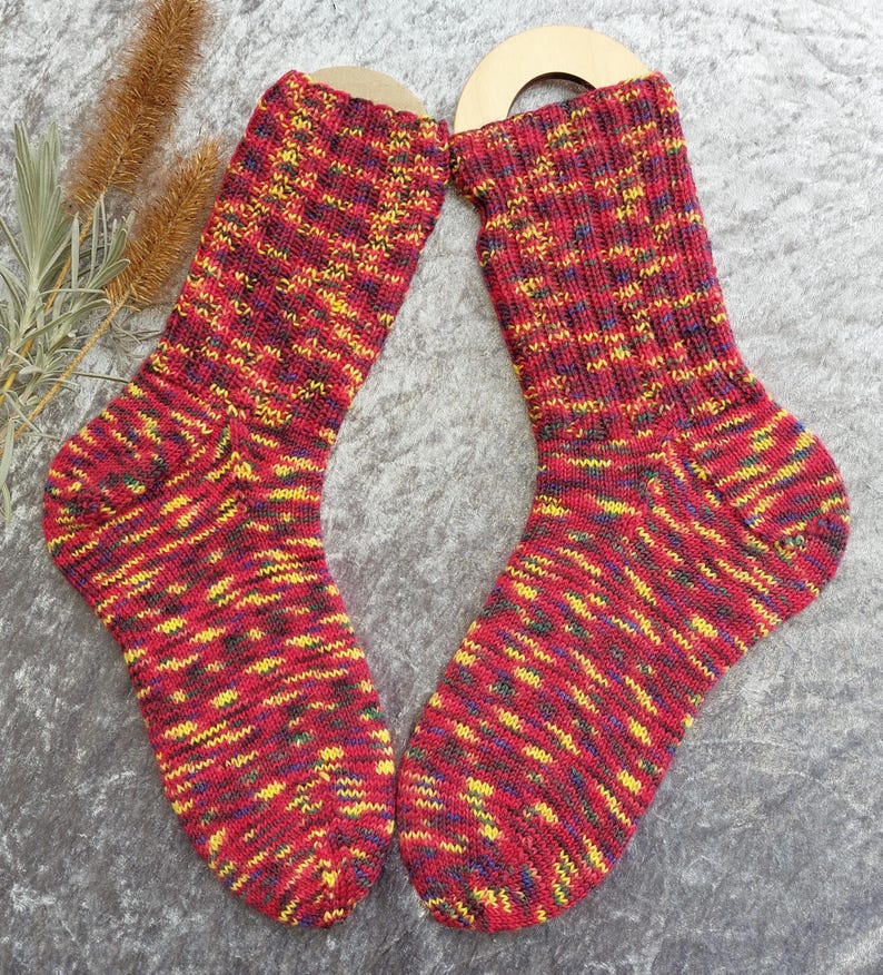 May include: A pair of colorful knitted socks. The socks are primarily red with yellow, blue, and green accents. The socks are displayed on wooden sock forms. The socks have a ribbed cuff and a patterned design throughout the body.