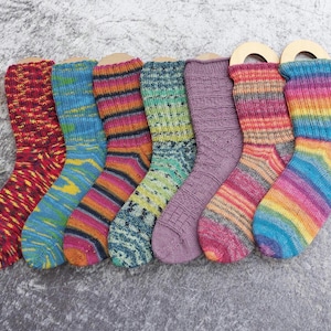 May include: A collection of seven colorful knitted socks, each with a unique pattern and color scheme. The socks are displayed on wooden forms, showcasing a variety of hues including red, blue, green, purple, and rainbow stripes. The socks appear to be made of wool or a similar yarn.