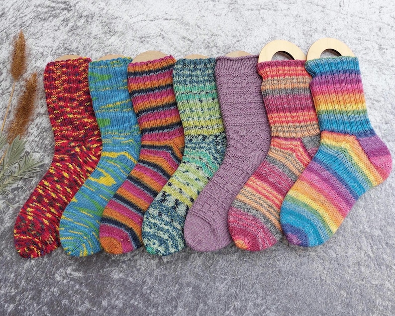 May include: A collection of seven colorful knitted socks, each with a unique pattern and color scheme. The socks are displayed on wooden forms, showcasing a variety of hues including red, blue, green, purple, and rainbow stripes. The socks appear to be made of wool or a similar yarn.