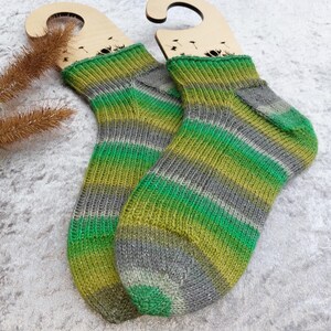 May include: A pair of hand-knitted socks with green, gray, and yellow stripes. The socks are displayed on wooden sock forms. The color pattern is repeated throughout the socks, with a ribbed cuff and a reinforced heel and toe.