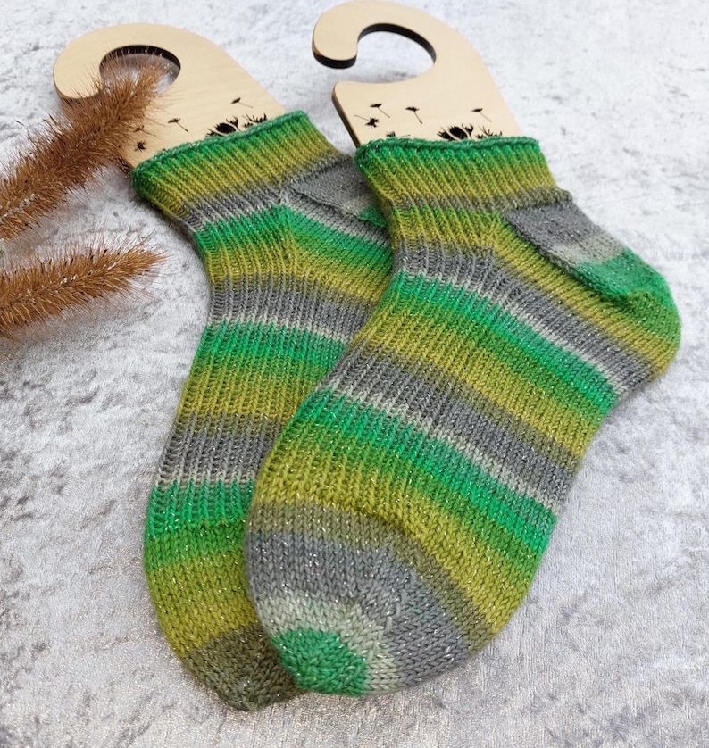 May include: A pair of hand-knitted socks with green, gray, and yellow stripes. The socks are displayed on wooden sock forms. The color pattern is repeated throughout the socks, with a ribbed cuff and a reinforced heel and toe.