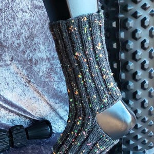 Könnte beinhalten: Dunkelgraue gestrickte Beinwärmer mit bunten Flecken, präsentiert auf einem schwarzen Schuhform. Die Beinwärmer haben eine gerippte Textur und sind so konzipiert, dass sie das untere Bein und den Knöchel bedecken. Ein schwarzes strukturiertes Objekt befindet sich im Hintergrund.
