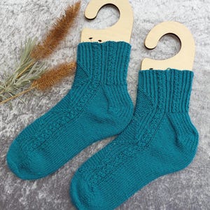 May include: A pair of teal knitted socks displayed on wooden hangers. The socks feature a textured pattern on the front and ribbed cuffs. The background is a textured gray surface, with dried plants on the left side.