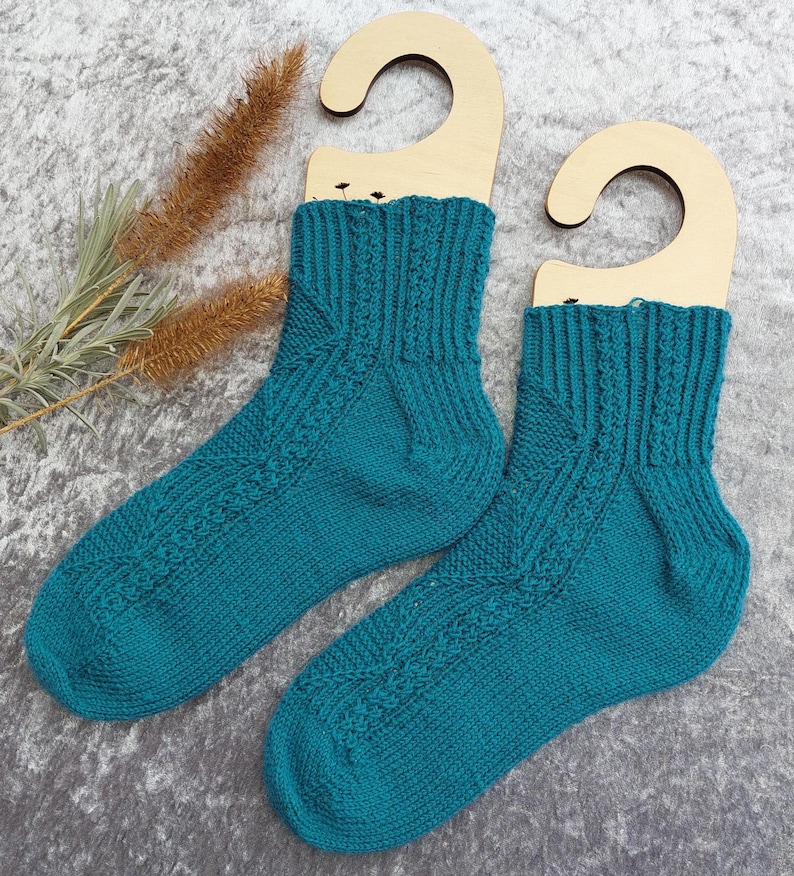 May include: A pair of teal knitted socks displayed on wooden hangers. The socks feature a textured pattern on the front and ribbed cuffs. The background is a textured gray surface, with dried plants on the left side.
